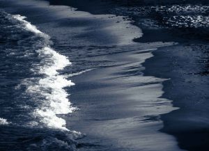 A white foaming wave cutting diagonally across dark wet sand, its edge sharp and luminous against the cool blue-grey of the shore.