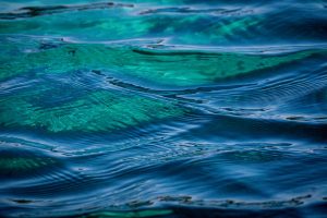 Deep blue-green water with gentle wave forms moving through the frame, patches of vivid emerald green light rising from the seabed below the translucent surface.