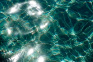 Intense white light refracting through shallow turquoise water onto a rocky seabed, the surface movement creating diagonal beams and bright pools across the green and teal bottom.