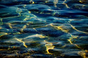 Deep blue water surface with rounded swells traced in iridescent rainbow edges and golden light seams.