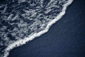 Close-up of a foaming wave arc curving across dark blue-grey sand, the white bubbles sharp and textured against the smooth shore.
