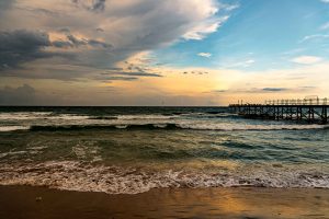 Waves roll onto a sandy beach at dusk under a dramatic sky split between storm clouds and golden light, a pier silhouetted on the right with birds perched along its railing.