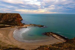 A sheltered crescent beach viewed from above, framed by red and ochre cliffs, the water a calm turquoise meeting the curved sandy shore under a cloudy sky at dusk.